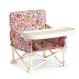The Paloma baby chair v2 is a portable booster seat for young children, featuring a colorful floral pattern, cream plastic tray, and foldable white metal legs—ideal for travel or dining.