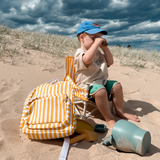 A young child in a blue cap sits on a dune under a cloudy sky, looking through binoculars. Next to them is a Brighton baby chair v2, sand toys, and a yellow-and-white striped backpack with a green water bottle.