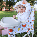 A baby in a fruit-patterned outfit and hat sits outdoors in the Clementine baby chair v2, holding a green spoon, with a grassy yard and trees in the background.